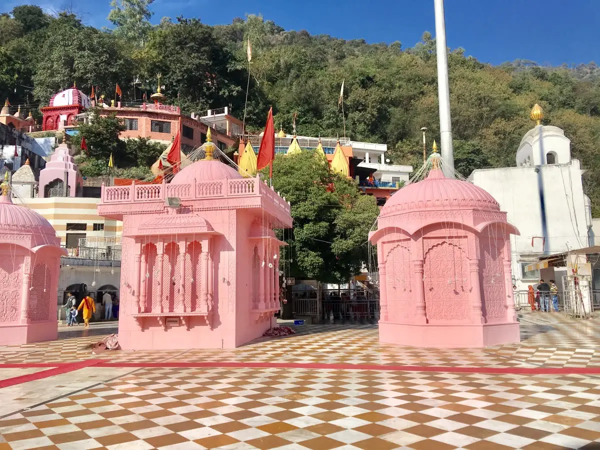 Pink pavilions and a tall flagpole on the checkered courtyard of Jwalamukhi temple below wooded hills